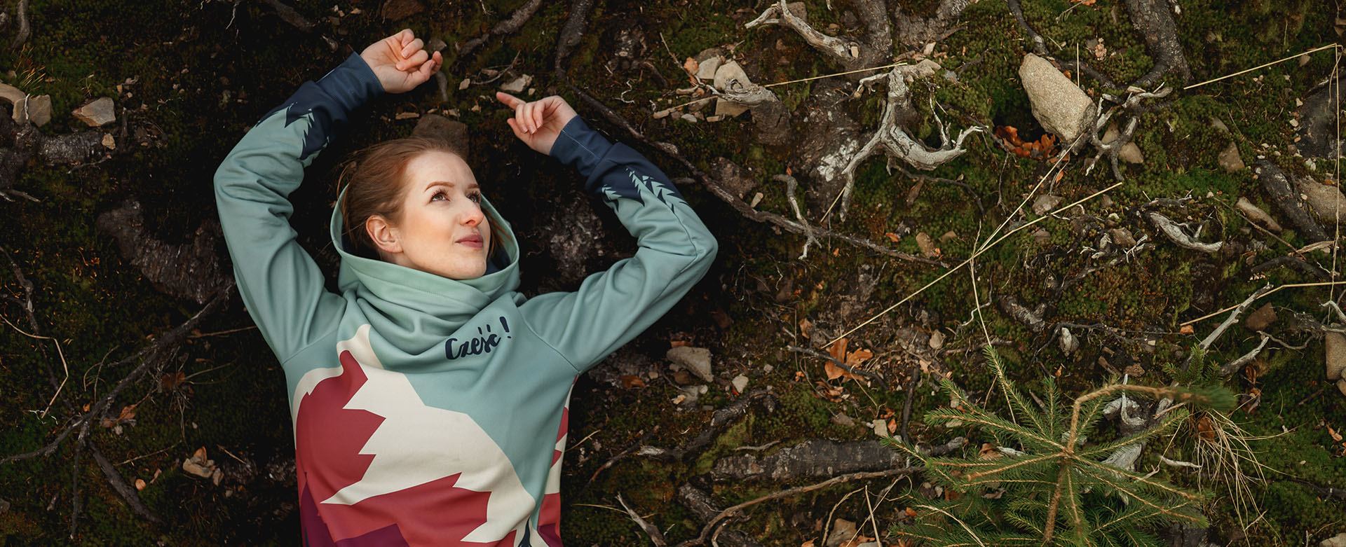 Woman lying on a mossy forest floor in a mountain-print hoodie with a wide hood